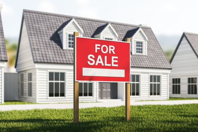Red “For Sale” sign in front of a white suburban house with a gray roof and green lawn