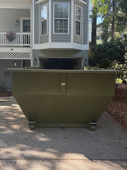 Large green dumpster on a driveway in front of a house with a bay window.
