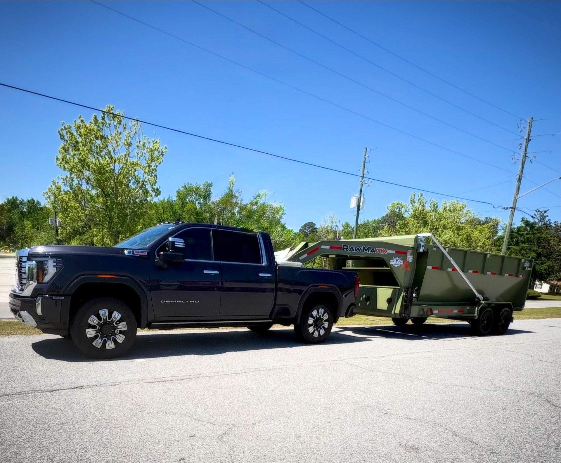 Black pickup truck towing a green dump trailer on a sunny roadside under a clear blue sky