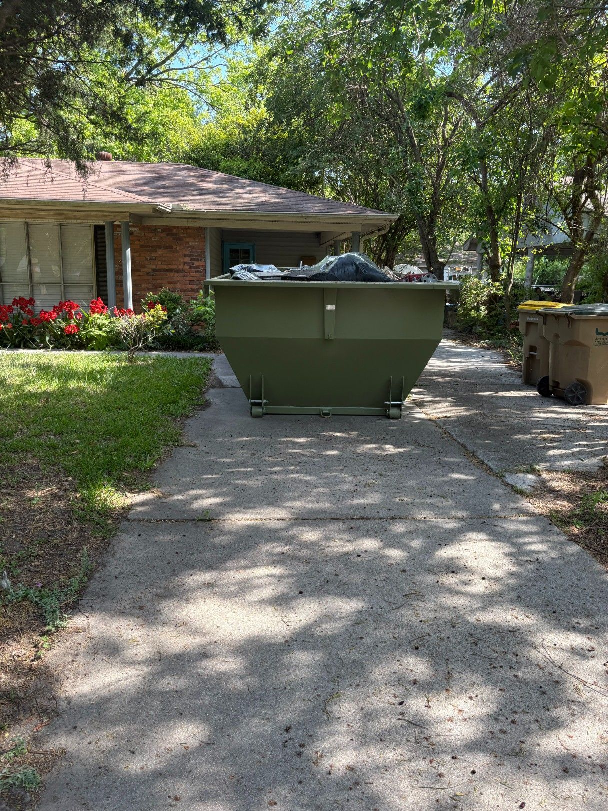 Green dumpster on a shaded driveway beside a house, with trees and a garage in the background