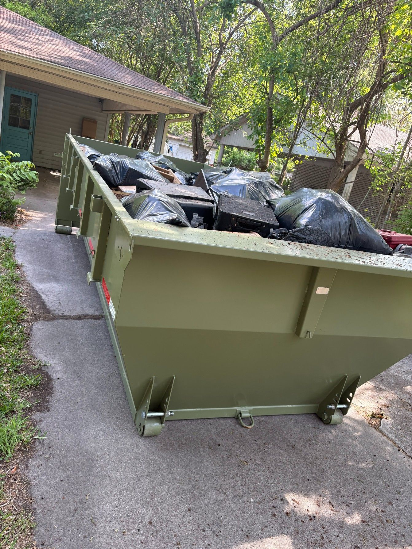Green dumpster filled with black trash bags on a driveway beside a house and trees