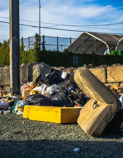 Overflowing trash pile beside a stone wall, with a yellow dumpster and loose garbage scattered on gravel.