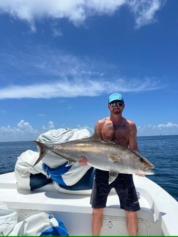 A man stands on a boat on a sunny day, holding a large fish he caught, with a blue sky and ocean in the background.