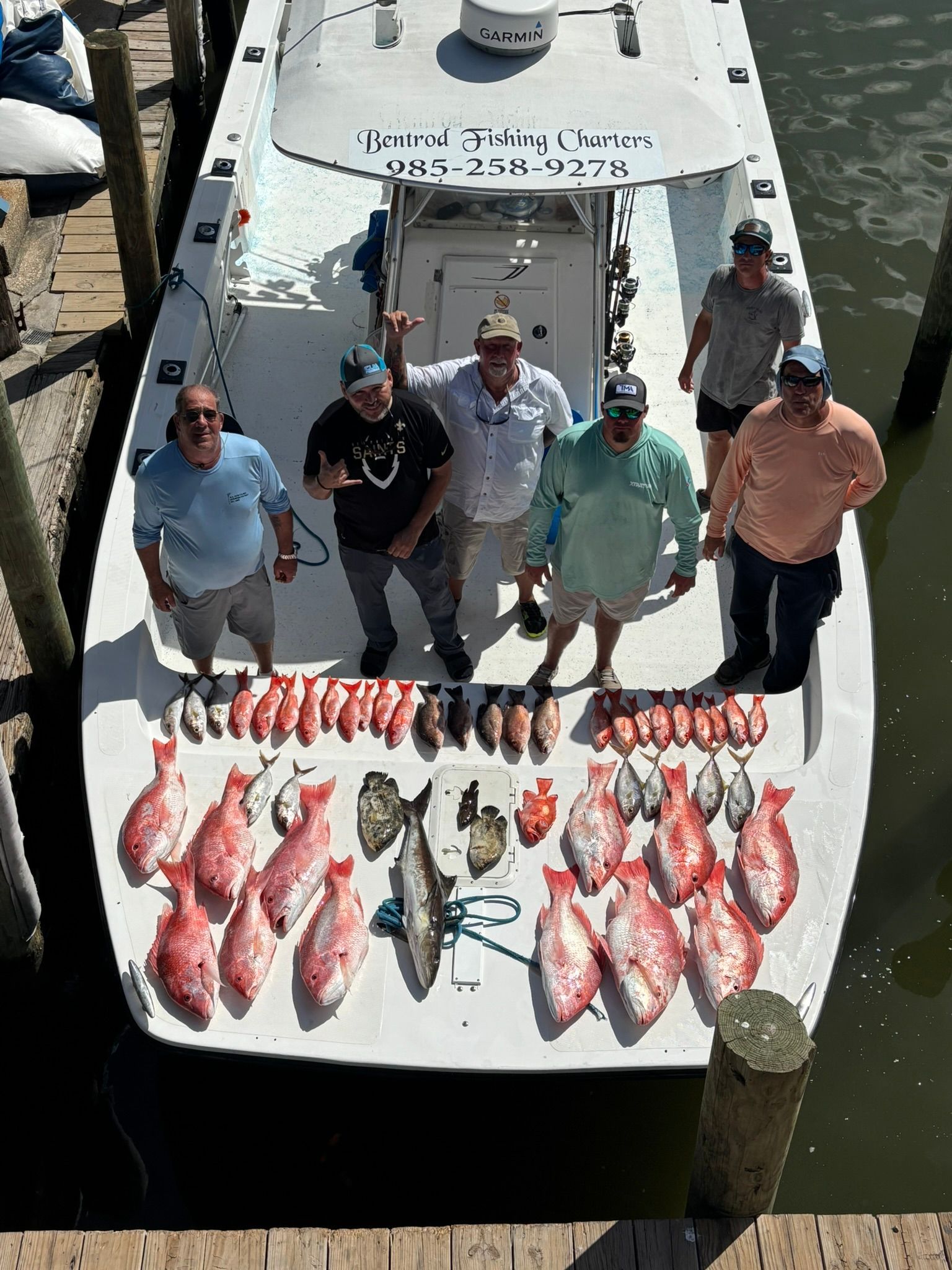 Six people stand on a boat deck behind a large catch of red snapper fish at a wooden dock.