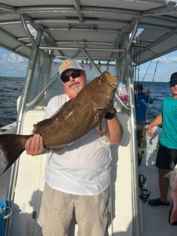 A person on a boat holding a large, brown fish caught while deep-sea fishing under a blue, sunny sky.