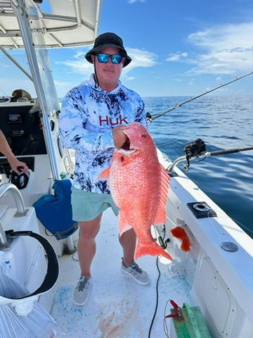A person wearing a sun hat and sunglasses stands on a boat holding a large red snapper fish.
