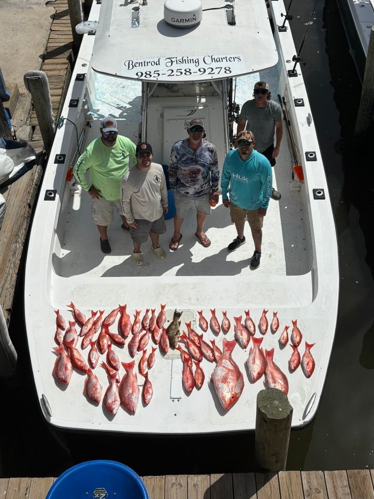 Five people stand on a boat deck covered with a large catch of red fish, docked at a pier.