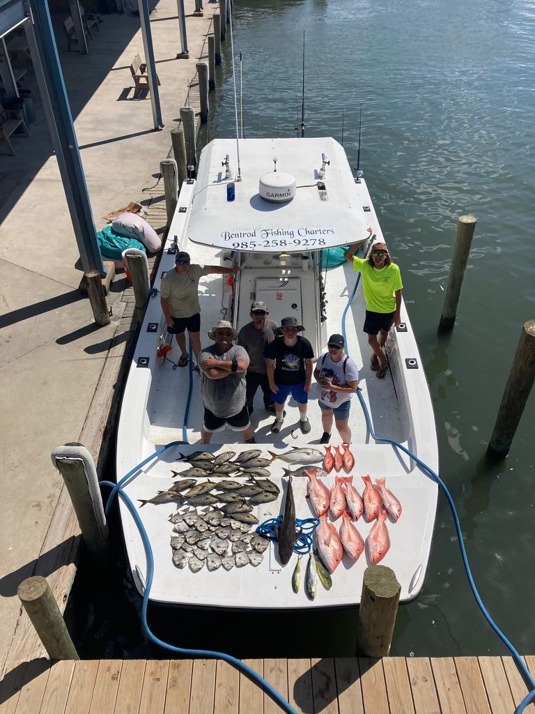 Six people stand on a white boat at a dock next to a large collection of freshly caught fish laid out on the deck.