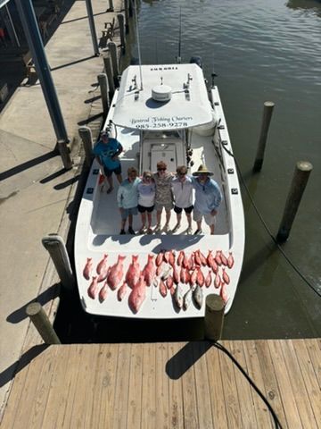 A group of people stand on a boat at a dock with a large catch of fish spread out on the deck.