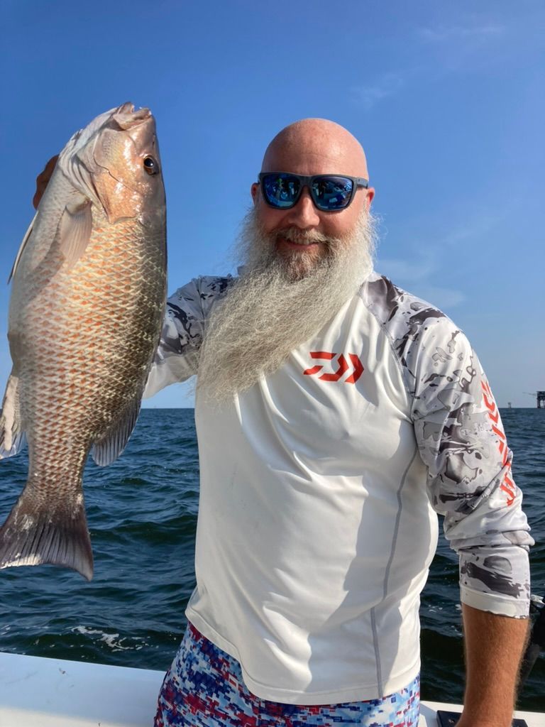 A person with a long white beard and sunglasses holds a large fish while standing on a boat against a blue sky.