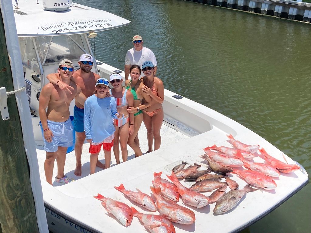 A group of people poses on a boat deck next to a large catch of fresh red snapper fish at a marina.