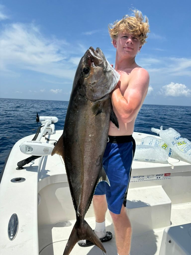 A person holding a large, dark-colored fish on a boat in the ocean under a clear blue sky.