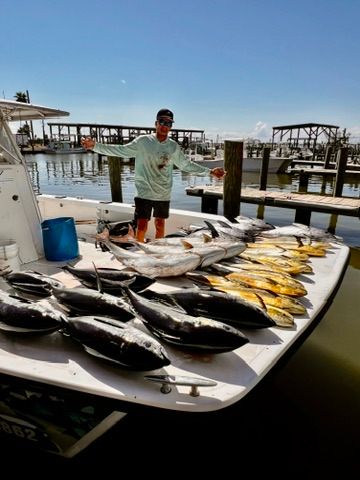 A person stands on a boat dock showing off a large catch of multiple tuna and mahi-mahi fish arranged on the boat deck.