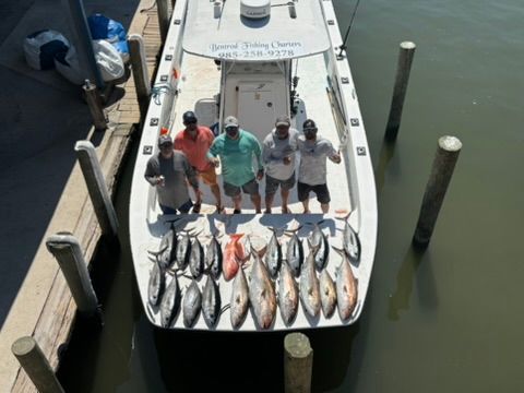 Five people stand on a boat docked at a pier, showcasing a large collection of fresh fish caught on the deck.