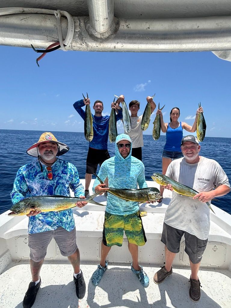 A group of six people on a boat deck hold freshly caught mahi-mahi fish under a bright blue sky.