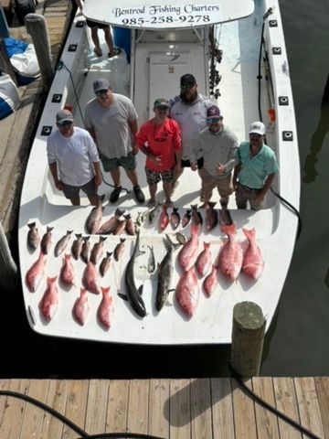 Six people stand on a charter fishing boat, looking down at a large catch of snapper and other fish spread on the deck.