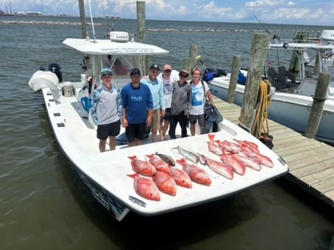 Six people stand on a boat at a dock next to a catch of red snapper arranged on the deck.