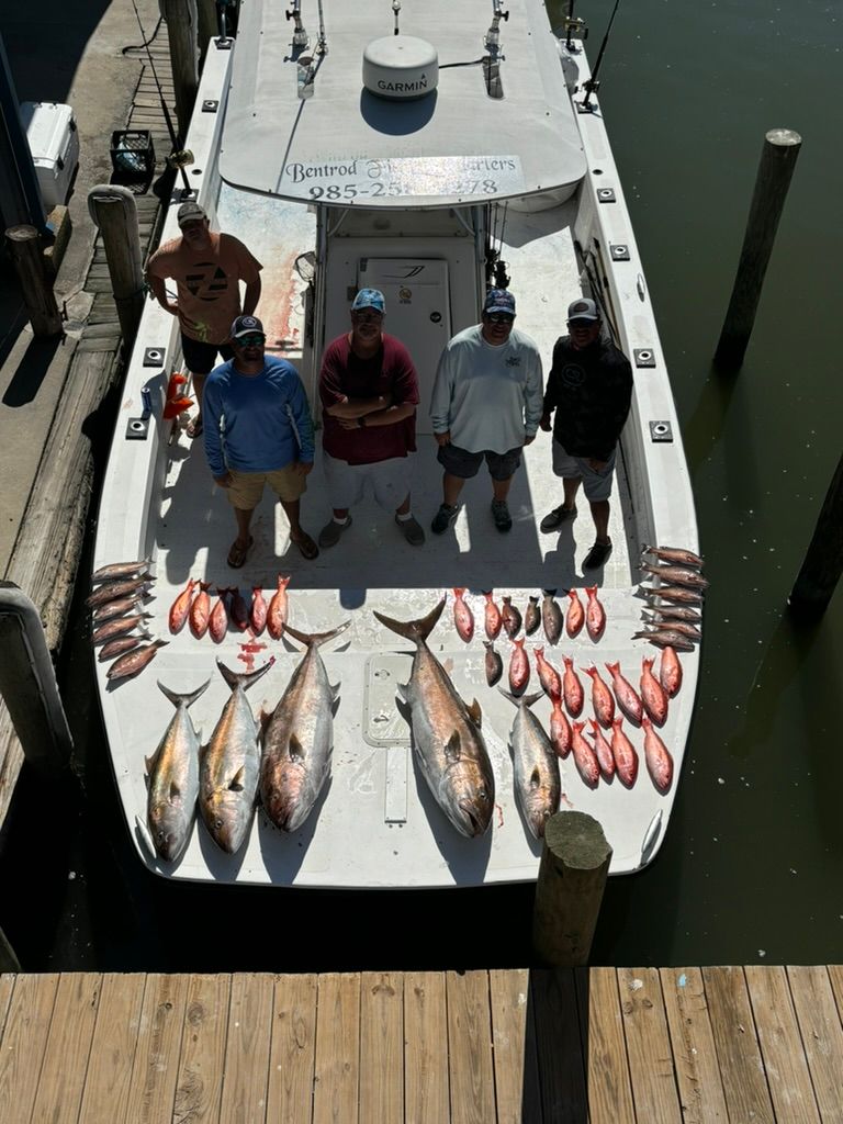 Five people stand on a boat deck next to a large haul of caught fish, docked at a pier.