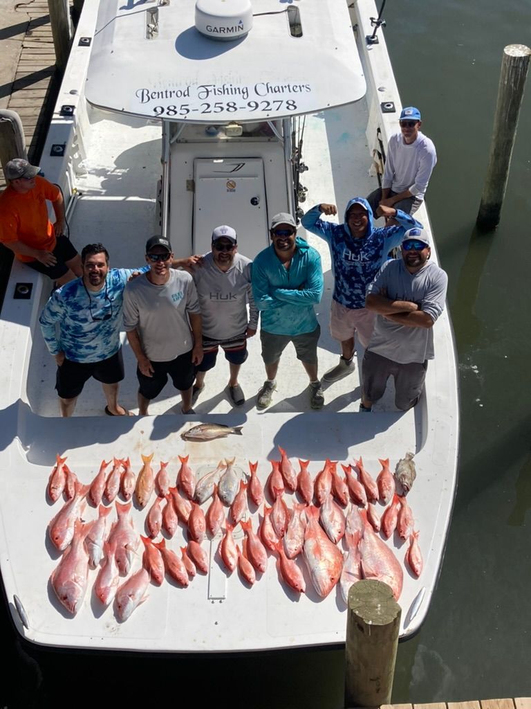 A group of people standing on a boat deck behind a large catch of red fish, with water and dock pilings visible nearby.