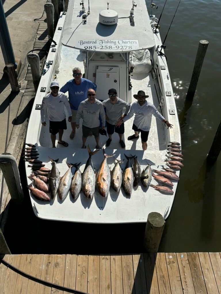 Five people stand on a white fishing boat docked at a pier, showcasing a large catch of fish spread across the deck.