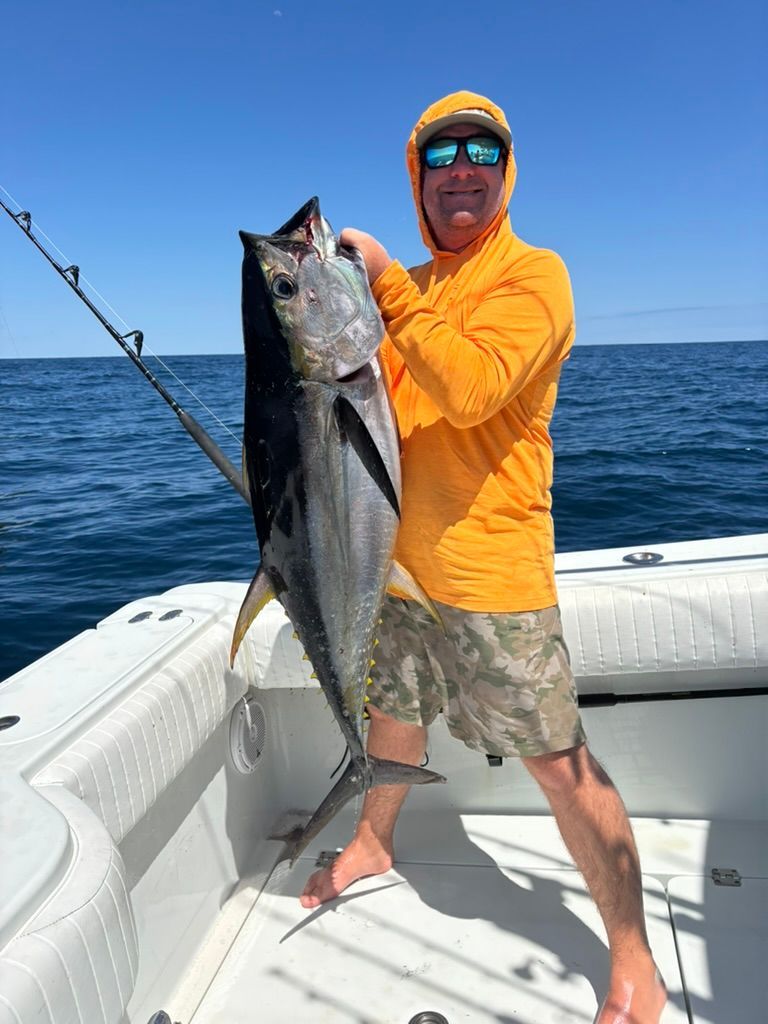 A person in an orange hooded shirt and camo shorts holds a large, silver tuna on a boat against a clear blue sky.