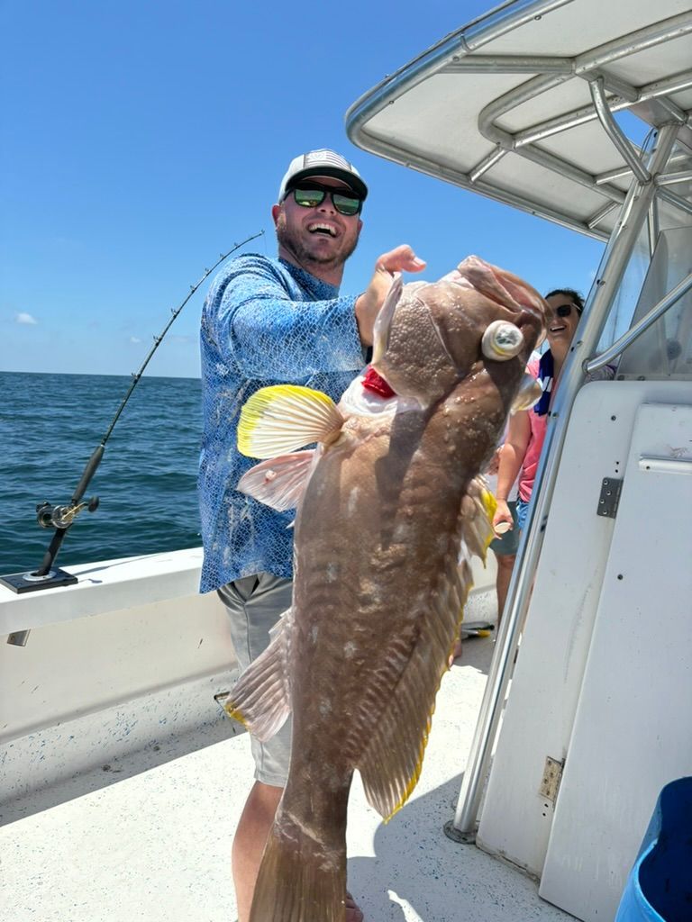 A person on a boat holding a large, brown grouper fish while smiling, with a second person visible in the background.