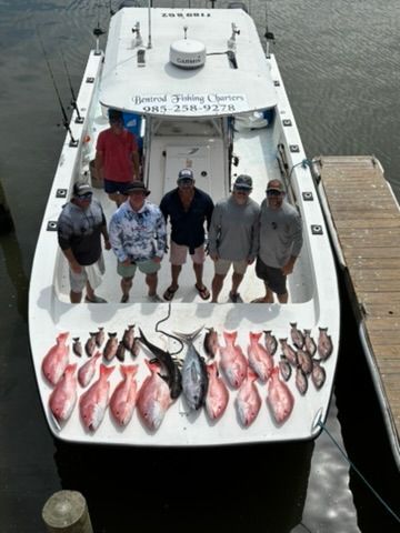 Six people stand on a boat docked at a pier, surrounded by a large catch of fresh fish laid out on the deck.