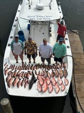 Five people stand on a charter fishing boat, behind a large catch of snapper fish arranged on the deck by a wooden pier.