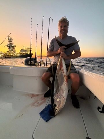 A person on a boat at sunset, holding a large tuna fish, with an industrial ship visible in the background.
