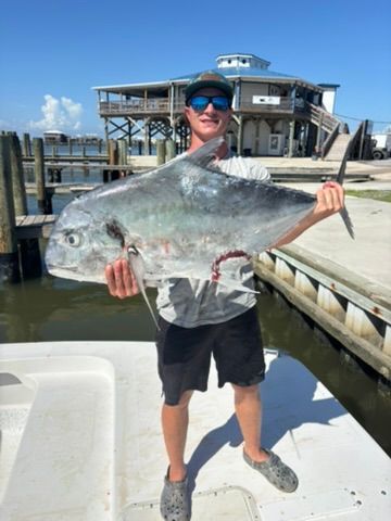 A person wearing sunglasses and a cap holds a large silver fish while standing on a boat dock near a waterfront building.