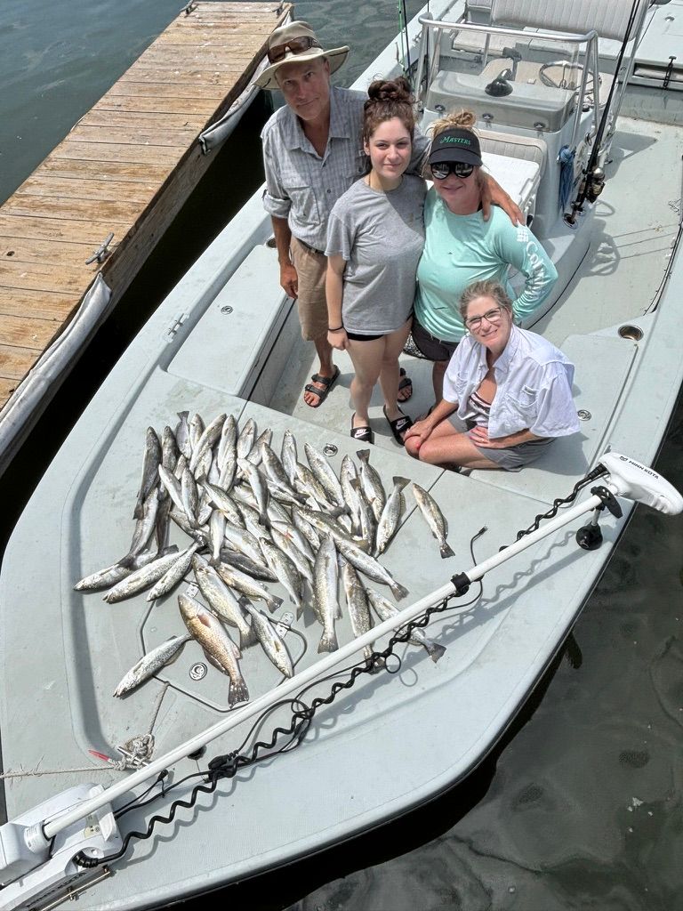 Four people pose on a boat deck next to a large catch of fish on the boat floor near a dock.