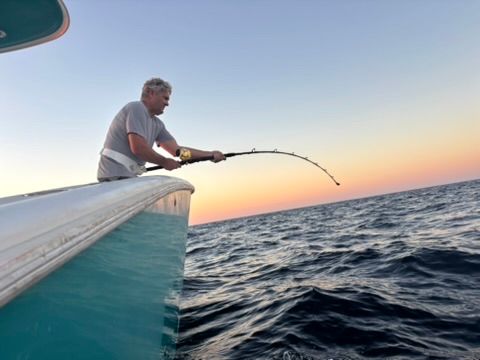 A person leans over the side of a boat at sunset, holding a fishing rod bent by a catch in the dark ocean water.