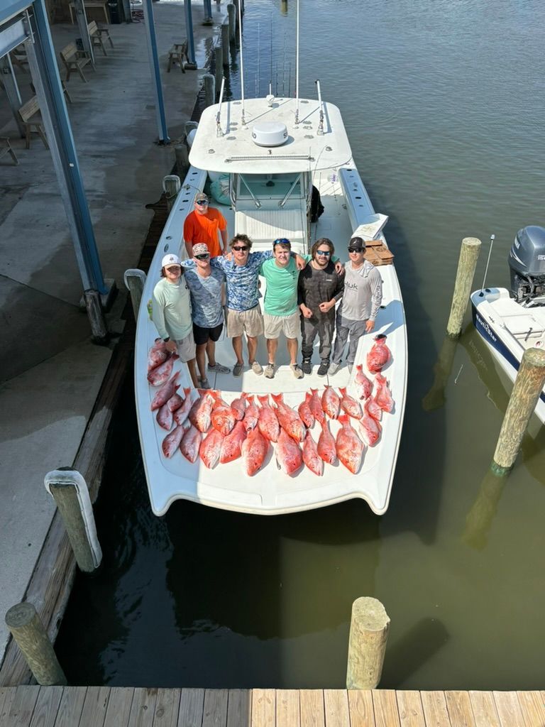 A group of people standing on a boat deck filled with red snapper fish at a dock.