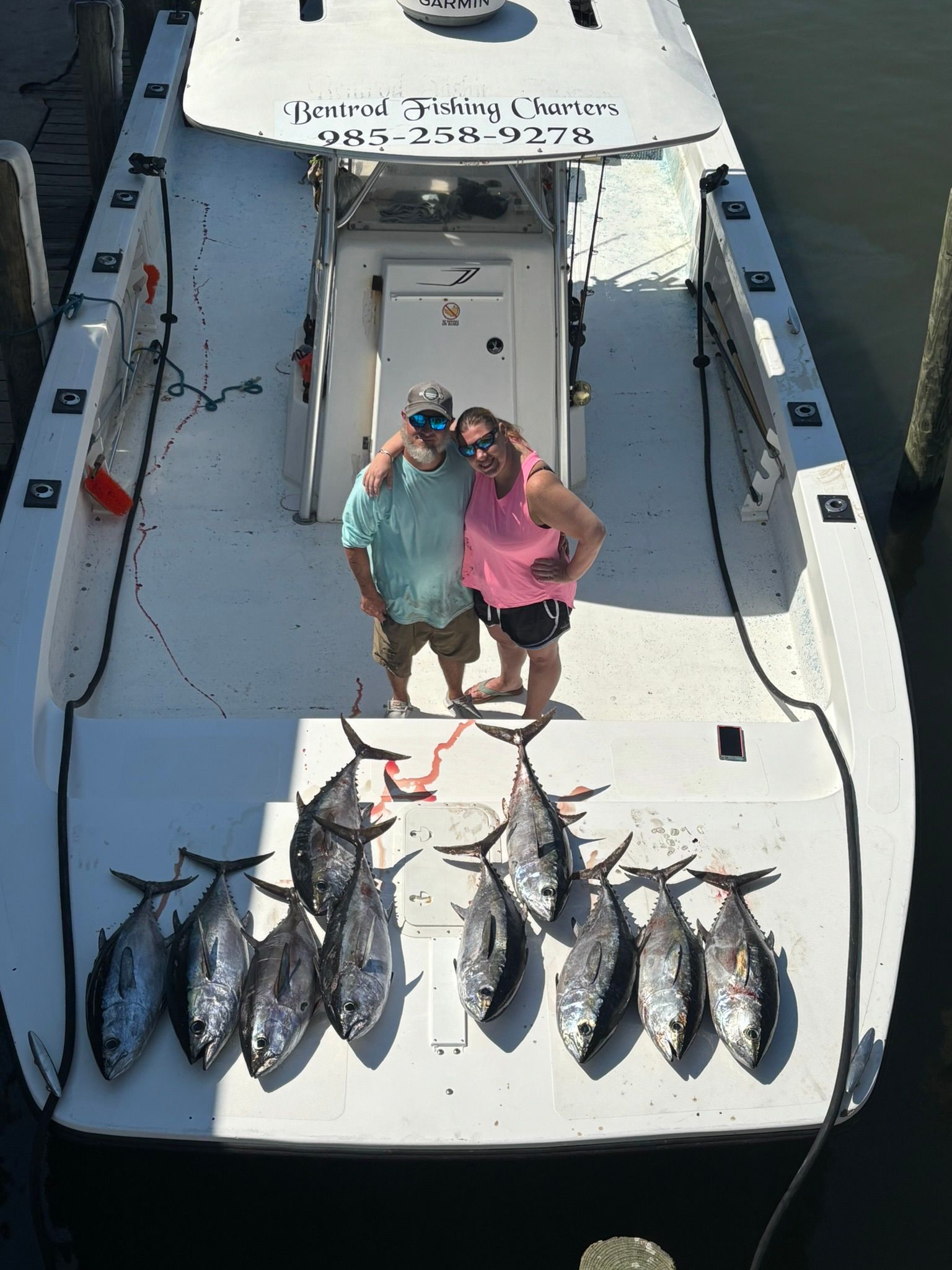 Two people stand on a boat deck next to a catch of ten tuna, with fishing equipment visible on the deck and boat exterior.