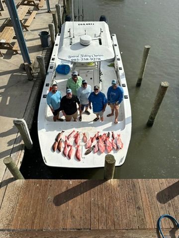 A group of five people stand on a white boat docked at a pier, displaying their catch of red snapper on the deck.