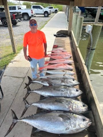 A person in an orange shirt stands next to a wooden dock displaying a catch of several tuna and snapper fish.