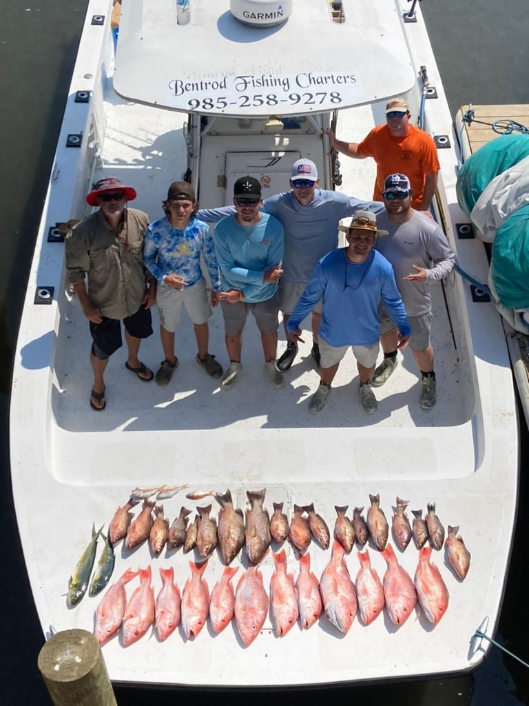 Group of eight people stand on a boat deck behind a large catch of fresh fish, with boat signage visible overhead.