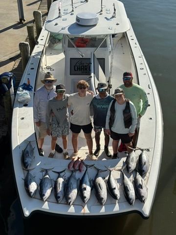 Six people stand on a boat deck surrounded by their catch of ten tuna fish, docked at a pier on a sunny day.