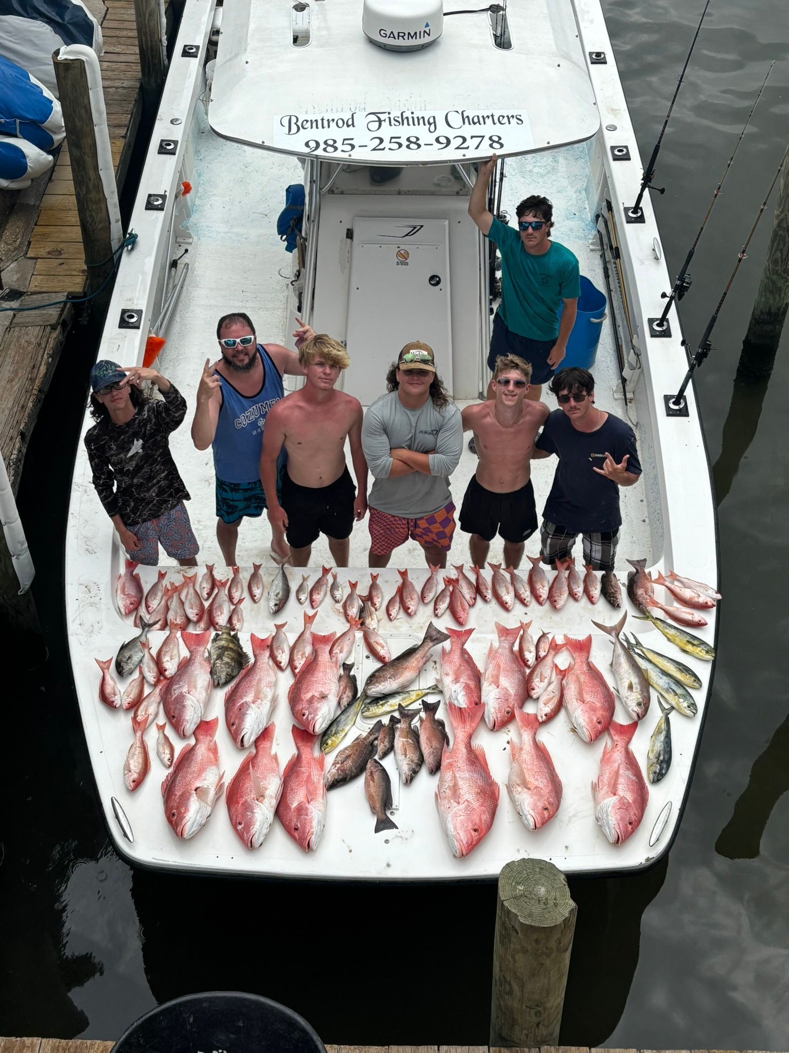 A group of seven people on a boat posing over a large catch of snapper and other fish, docked at a pier.