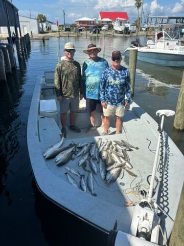 Three people stand on a boat docked at a marina with a catch of fish spread on the deck.