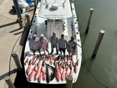 Five people stand on a fishing boat at a dock next to a large haul of caught red snapper and two large cobia fish.