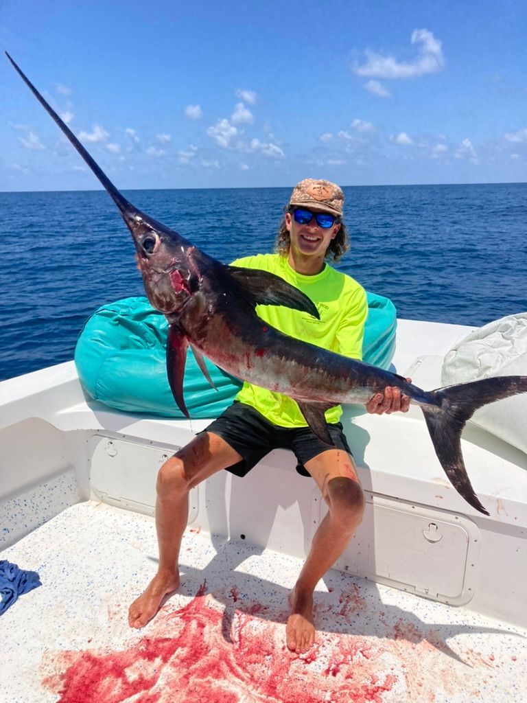 A person in a bright yellow shirt and sunglasses holds a large swordfish on a boat deck with an ocean background.