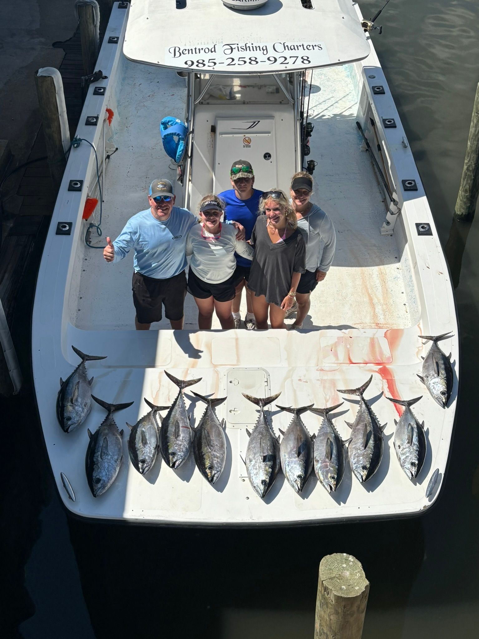A group poses on a boat deck behind a catch of twelve tuna, with water visible around the boat.