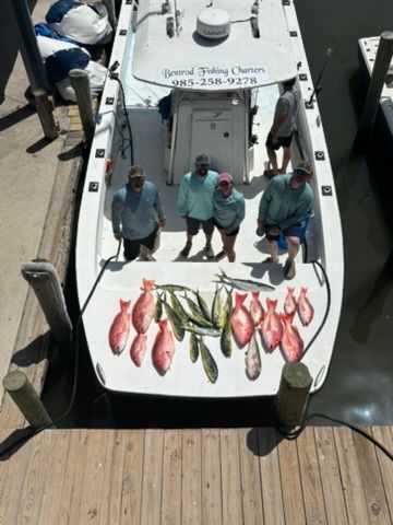 Four people stand on a boat docked at a pier, displaying their catch of red snapper and mahi-mahi on the deck.