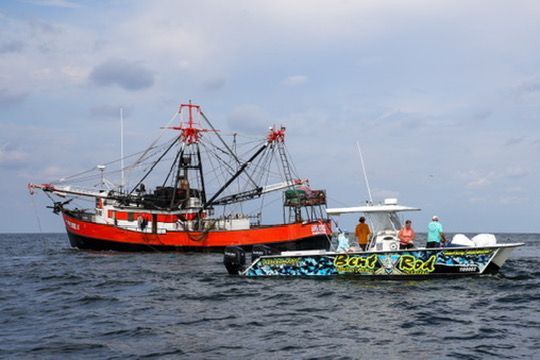 A red shrimp trawler floats on the water next to a smaller, patterned recreational boat with four people aboard.