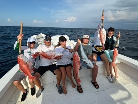 A group of six people on a boat at sea holding freshly caught red snapper and a shark while smiling for a photo.