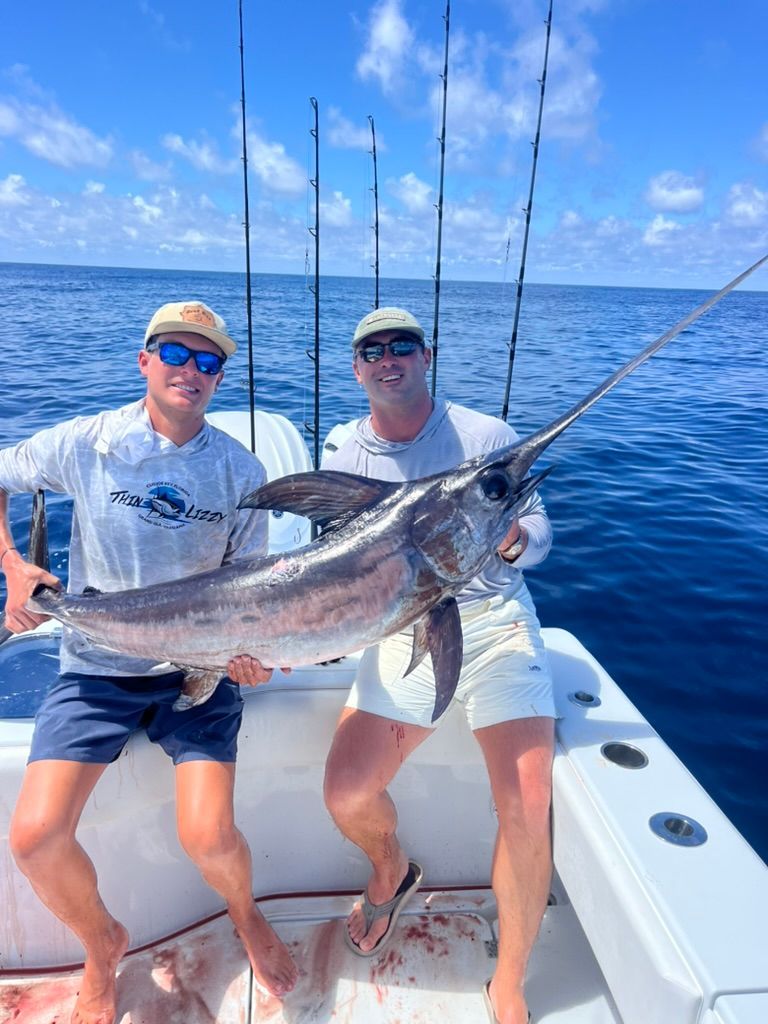 Two people stand on a boat deck, smiling and holding a large billfish caught in the open ocean.