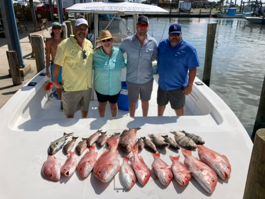 Four people pose on a boat deck behind a large catch of red snapper and other fish, docked at a marina.