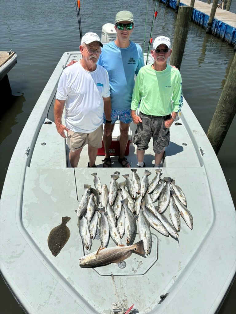 Three people stand on a boat with a catch of fish laid out on the deck at a dock.