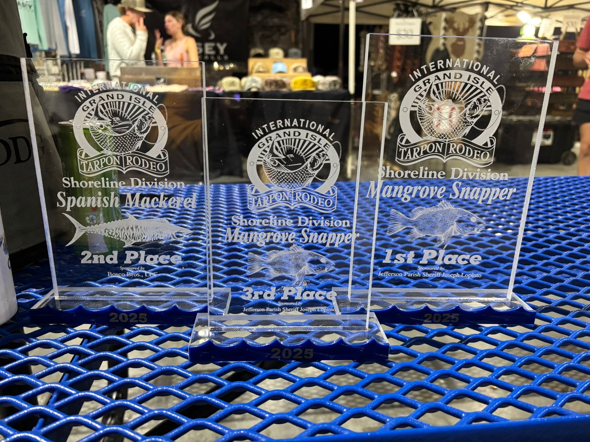 Three clear acrylic fishing tournament trophies for 2025 displayed on a blue metal table.